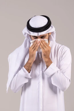 The need for treatment and medicine, the necessity of going to the doctor, feeling ill and having a cold, nasal inflammation and allergic sinuses, a portrait of an Emirati Gulf Arab man wearing a kandura and ghutrah holding a tissue to his face with sneezing gestures, white background.