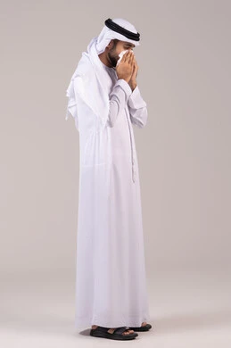 Feeling sick and having a cold, nasal inflammation and allergic sinuses, the need for treatment and medication, a side portrait of an Emirati Gulf Arab man wearing a kandura and ghutrah holding a tissue to his face with sneezing gestures, full body, white background.