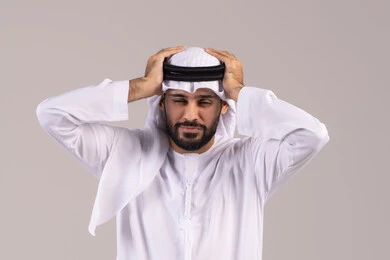 الشعور بالصداع وألم الرأس, الاحساس بالشقيقية والصداع النصفي, الاجهاد والضغط النفسي, بورتريه مقرب لرجل عربي خليجي اماراتي يرتدي الكندورة والغترة يضع يديه على رأسه ويغمض عينيه بإيماءات الألم الشديد, الإرهاق وفقدان التركيز, خلفية بيضاء