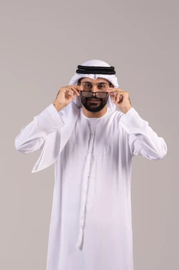 مفهوم تعديل وتحسين الرؤية، الاناقة والاهتمام بالمظهر الخارجي، بورتريه لرجل عربي خليجي اماراتي يرتدي الكندورة والغترة يمسك النظاره الطبيه، ارتداء النظارات الخاصة بضعف النظر، خلفية بيضاء