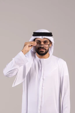 The concept of correcting and improving vision, elegance, and attention to appearance, a portrait of an Arab Gulf Emirati man wearing a kandura and ghutrah holding prescription glasses, wearing glasses for vision impairment, white background.