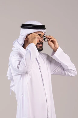 The concept of correcting and improving vision, elegance, and attention to appearance, a portrait of an Arab Gulf Emirati man wearing a kandura and ghutrah holding prescription glasses, wearing glasses for vision impairment, white background.