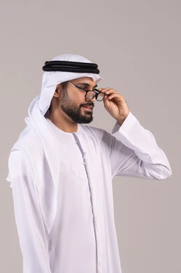 The concept of correcting and improving vision, elegance, and attention to appearance, a portrait of an Arab Gulf Emirati man wearing a kandura and ghutrah holding prescription glasses, wearing glasses for vision impairment, white background.