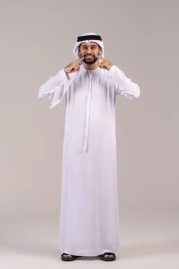 الحاجة إلى علاج سني، الشعور بآلام الفك والأسنان، الاشارة بإصبع السبابة إلى الأسنان، الوقوف باستقامة والنظر إلى الكاميرا بإيماءات التألم، مفهوم الاهتمام بالمظهر الخارجي، بورتريه لرجل عربي خليجي اماراتي يرتدي الكندورة والغترة ، الطول الكامل للجسم، خلفية بيضاء