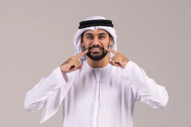 الوقوف باستقامة والنظر إلى الكاميرا بإيماءات التألم، الحاجة إلى علاج سني، الشعور بآلام الفك والأسنان، الاشارة بإصبع السبابة إلى الأسنان، مفهوم الاهتمام بالمظهر الخارجي، بورتريه مقرب لرجل عربي خليجي اماراتي يرتدي الكندورة والغترة ، خلفية بيضاء