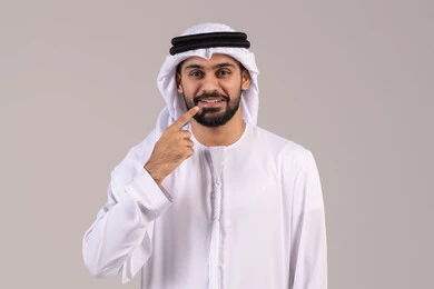 Standing upright and looking at the camera with gestures of pain, the need for dental treatment, feeling pain in the jaw and teeth, pointing with the index finger to the teeth, the concept of caring about appearance, a close-up portrait of an Emirati Gulf Arab man wearing a kandura and ghutrah, white background. Standing upright and looking at the camera with gestures of pain, the need for dental treatment, feeling pain in the jaw and teeth, pointing with the index finger to the teeth, the concept of caring about appearance, a close-up portrait of an Emirati Gulf Arab man wearing a kandura and ghutrah, white background.