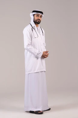 مفهوم الطب والرعاية الصحية، العمل في القطاع الصحي في الامارات, الوقوف باستقامة بإيماءات الثقة بالنفس، بورتريه لطبيب عربي خليجي اماراتي يرتدي الكندورة والغترة والمعطف الطبي يضع السماعة الطبية ينظر إلى شيء ما في الاعلى، الجسم بالطول الكامل، خلفية بيضاء