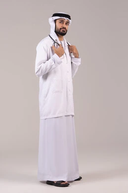 مفهوم الطب والرعاية الصحية، العمل في القطاع الصحي في الامارات, الوقوف باستقامة بإيماءات الثقة بالنفس، بورتريه لطبيب عربي خليجي اماراتي يرتدي الكندورة والغترة والمعطف الطبي يضع السماعة الطبية ينظر إلى شيء ما في الاعلى، الجسم بالطول الكامل، خلفية بيضاء