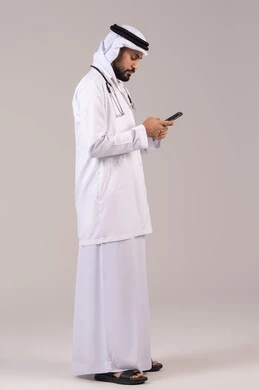 Meeting the demands of telehealth using modern technologies, showcasing medical services and applications via phone, a portrait of an Arab Gulf Emirati doctor wearing a kandura, ghutrah, and medical coat, holding a stethoscope and a mobile phone in hand, full-body, white background.