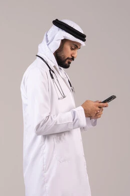 عرض خدمات و تطبيقات طبية عبر الهاتف، تلبية مطالب الرعاية الصحية عن بعد باستخدام التقنيات الحديثه، بورتريه طبيب عربي خليجي اماراتي يرتدي الكندورة والغترة والمعطف الطبي ويضع السماعة الطبية يحمل بيده الهاتف المحمول، خلفية بيضاء
