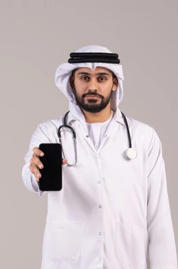 عرض شاشة سوداء فارغة عبر الجوال، خدمات و تطبيقات طبية عبر الهاتف، تلبية مطالب الرعاية الصحية عن بعد باستخدام التقنيات الحديثه، بورتريه طبيب عربي خليجي اماراتي يرتدي الكندورة والغترة والمعطف الطبي ويضع السماعة الطبية يحمل بيده الهاتف المحمول، خلفية بيضاء