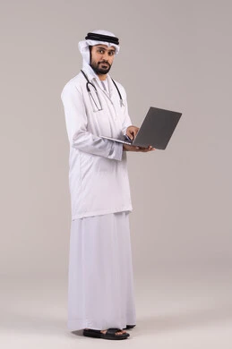 استخدام الأجهزة التقنية الحديثة في الطب، بورتريه لطبيب عربي خليجي اماراتي يرتدي الكندورة والغترة والمعطف الأبيض يحمل بيده الحاسوب المحمول ينظر إلى الكاميرا، تقديم الاستشارات الطبية عن طريق الإنترنت، العمل في القطاع الصحي، الجسم بالطول الكامل، خلفية بيضاء