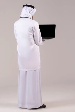 Providing medical consultations via the internet, using modern technological devices in medicine, a side portrait of an Emirati Gulf Arab doctor wearing a kandura, ghutrah, and white coat holding a laptop in his hand looking at the camera, working in the healthcare sector, full body length, white background.