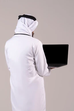 Working in the healthcare sector, providing medical consultations via the internet, using modern technological devices in medicine, a portrait from behind of an Arab Gulf Emirati doctor wearing a kandura, ghutrah, and white coat holding a laptop in his hand, white background.