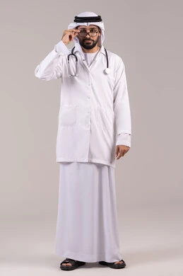تقديم الخدمات العلاجية للمرضى, الوقوف باستقامة بإيماءات الثقة بالنفس، العمل في القطاع الصحي في الامارات، بورتريه لطبيب عربي خليجي اماراتي يرتدي الكندورة والغترة والمعطف الطبي يضع السماعة والنظارة الطبية ينظر إلى الكاميرا، الجسم بالطول الكامل، خلفية بيضاء