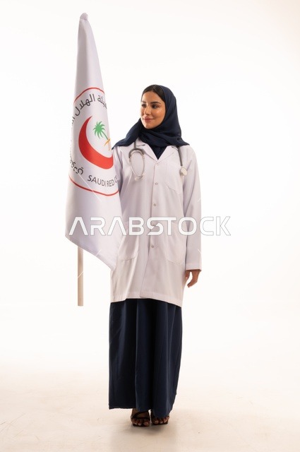 علم الهلال السعودي، مفهوم الطب والرعاية الصحية ، بورتريه لطبيبة عربية خليجية سعودية ترتدي العباءة والمئزر الأبيض تمسك بيديها علم عليه شعارالهلال الأحمر، شابة سعودية تنظر إلى الكاميرا بتعابير الحزم والجدية, خلفية بيضاء