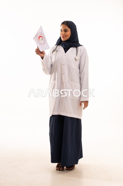 {مفهوم الطب والرعاية الصحية ، بورتريه لطبيبة عربية خليجية سعودية ترتدي العباءة والمئزر الأبيض تمسك بيديها علم الهلال الأحمر، شابة سعودية تنظر إلى الكاميرا بتعابير الحزم والجدية، العروض والإعلانات التسويقية, خلفية بيضاء