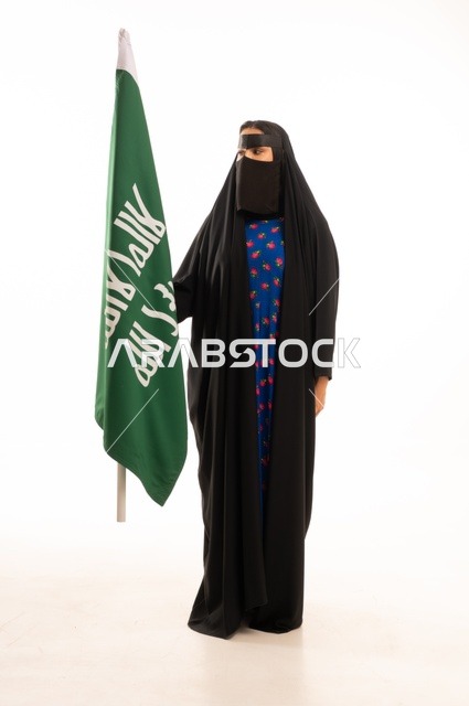 ارتداء الأزياء الشعبية في المناسبات الوطنية, ذكرى يوم بدينا 1727 م، تأسيس الدولة السعودية الأولى 22 فبراير، بورتريه لامرأة عربية خليجية سعودية ترتدي البرقع والعباءة تمسك راية وشعار يوم التأسيس بتعابير الفخر والاعتزاز, الطول الكامل للجسم، خلفية بيضاء