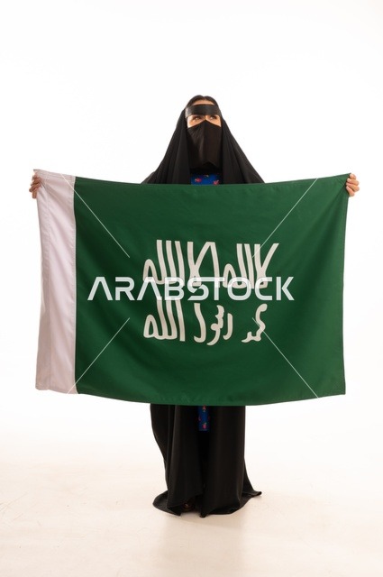 Wearing traditional attire on national occasions, commemorating the Day of Foundation 1727 AD, the establishment of the first Saudi state on February 22, a portrait of an Arab Gulf Saudi woman wearing a burqa and abaya holding the flag and emblem of the Day of Foundation with expressions of pride and honor, full body length, white background.