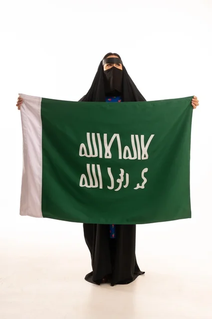 ارتداء الأزياء الشعبية في المناسبات الوطنية, ذكرى يوم بدينا 1727 م، تأسيس الدولة السعودية الأولى 22 فبراير، بورتريه لامرأة عربية خليجية سعودية ترتدي البرقع والعباءة تمسك راية وشعار يوم التأسيس بتعابير الفخر والاعتزاز, الطول الكامل للجسم، خلفية بيضاء