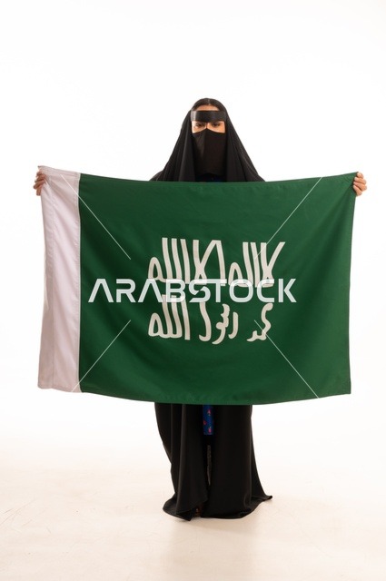ارتداء الأزياء الشعبية في المناسبات الوطنية, ذكرى يوم بدينا 1727 م، تأسيس الدولة السعودية الأولى 22 فبراير، بورتريه لامرأة عربية خليجية سعودية ترتدي البرقع والعباءة تمسك راية وشعار يوم التأسيس بتعابير الفخر والاعتزاز, الطول الكامل للجسم، خلفية بيضاء