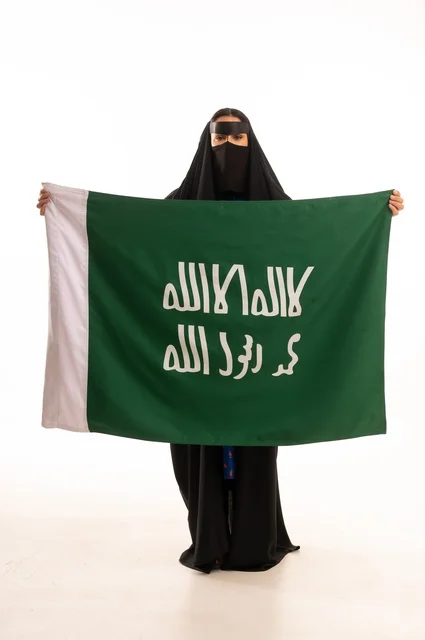 ارتداء الأزياء الشعبية في المناسبات الوطنية, ذكرى يوم بدينا 1727 م، تأسيس الدولة السعودية الأولى 22 فبراير، بورتريه لامرأة عربية خليجية سعودية ترتدي البرقع والعباءة تمسك راية وشعار يوم التأسيس بتعابير الفخر والاعتزاز, الطول الكامل للجسم، خلفية بيضاء