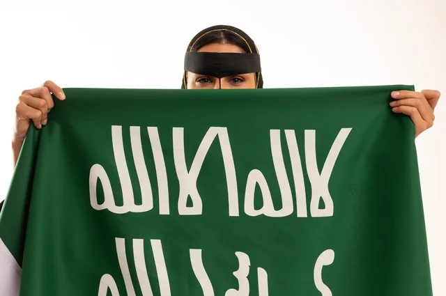 ذكرى يوم بدينا 1727 م، تأسيس الدولة السعودية الأولى 22 فبراير، ارتداء الأزياء الشعبية في المناسبات الوطنية, بورتريه مقرب لإمرأة عربية خليجية سعودية ترتدي البرقع تمسك راية وشعار يوم التأسيس بتعابير الفخر والاعتزاز, خلفية بيضاء