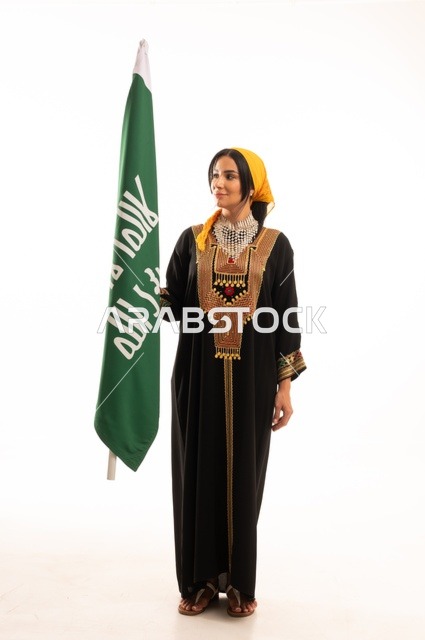 الاحتفال بالمناسبات والأعياد الوطنية الشعبية، ذكرى تأسيس الدولة السعودية الأولى 22 فبراير، يوم بدينا 1727 م، ارتداء أزياء المنطقة الوسطى، بورتريه لإمرأة عربية خليجية سعودية مبتسمة ترتدي الكرتة والهامة تحمل بيدها علم يوم التأسيس، الجسم بالطول الكامل، خلفية بيضاء