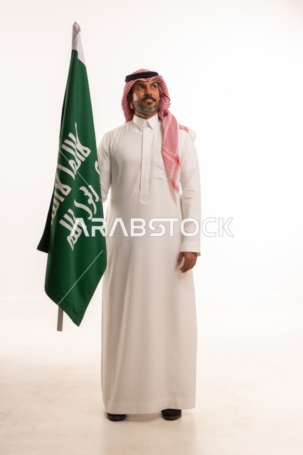 إحياء ذكرى اليوم الوطني السعودي 23 سبتمبر, إيماءات الفخر و السرور بالانتماء للوطن, بورتريه لرجل عربي خليجي سعودي مبتسم يرتدي الشماغ و الثوب التقليدي يحمل بيده علم يوم التأسيس، يوم بدينا 1727 م، الجسم بالطول الكامل، خلفية بيضاء