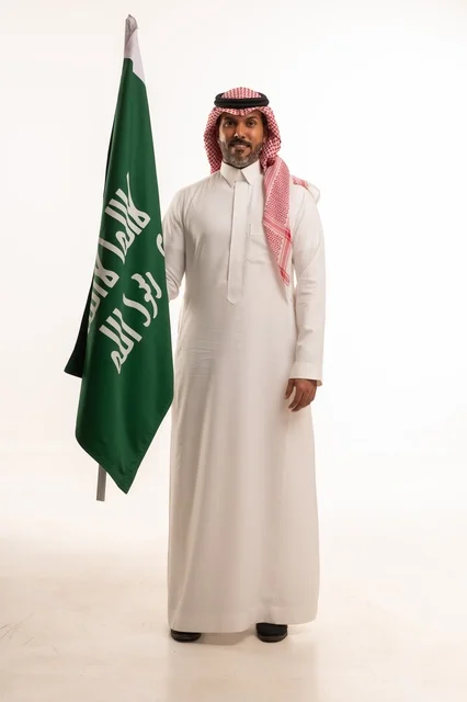 إحياء ذكرى اليوم الوطني السعودي 23 سبتمبر, إيماءات الفخر و السرور بالانتماء للوطن, بورتريه لرجل عربي خليجي سعودي مبتسم يرتدي الشماغ و الثوب التقليدي يحمل بيده علم يوم التأسيس، يوم بدينا 1727 م، الجسم بالطول الكامل، خلفية بيضاء