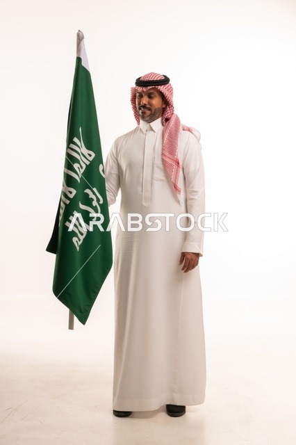 إحياء ذكرى اليوم الوطني السعودي 23 سبتمبر, إيماءات الفخر و السرور بالانتماء للوطن, بورتريه لرجل عربي خليجي سعودي مبتسم يرتدي الشماغ و الثوب التقليدي يحمل بيده علم يوم التأسيس، يوم بدينا 1727 م، الجسم بالطول الكامل، خلفية بيضاء