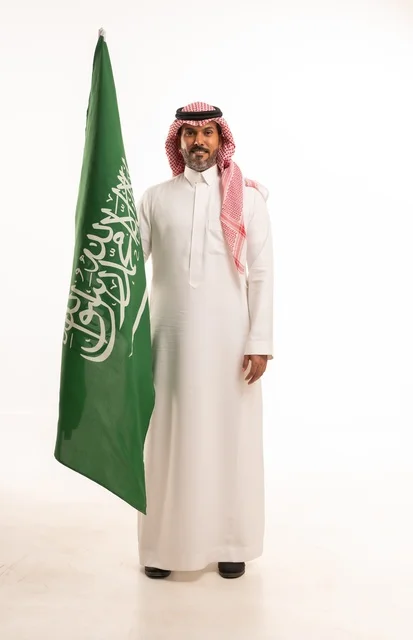 يوم العلم السعودي 11 مارس، إحياء ذكرى اليوم الوطني السعودي 23 سبتمبر, إيماءات الفخر و السرور بالانتماء للوطن, بورتريه لرجل عربي خليجي سعودي مبتسم يرتدي الشماغ و الثوب التقليدي يحمل بيده علم المملكة، يوم بدينا 1727 م، الجسم بالطول الكامل، خلفية بيضاء