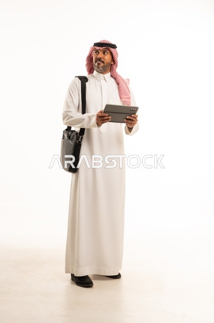 إنجاز مهام العمل عن بعد، التسوق الالكتروني وطلبات الاونلاين ، دردشة ومحادثات و تواصل اجتماعي ، اجراء اجتماع عمل عبر الانترنت، بورتريه لرجل عربي سعودي خليجي يرتدي الثوب والشماغ التقليدي يحمل حقيبة العمل ويستخدم التابليت المحمول، الجسم بالطول الكامل، خلفية بيضاء