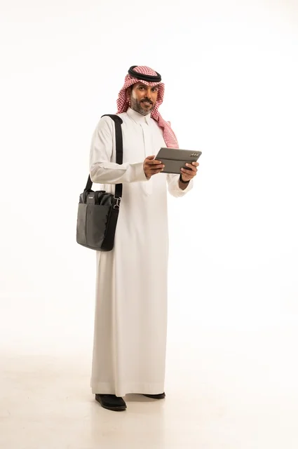 Completing remote work tasks, online shopping and orders, chatting and social communication, conducting a business meeting online, a portrait of an Arab Saudi Gulf man wearing traditional thobe and ghutrah, carrying a briefcase and using a tablet, full-body, white background.