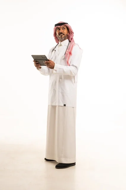 الاهتمام بالمظهر الخارجي، دمج التطور التقني في الحياة العملية، تقديم الفحص الطبي والخدمات العلاجية، بورتريه مقرب لطبيب عربي خليجي سعودي مبتسم يرتدي معطف الطبيب ويضع السماعات الطبية يحمل بيده جهاز لوحي يستخدمه لمراجعة حالات المرضى، الرعاية الصحية، خلفية بيضاء