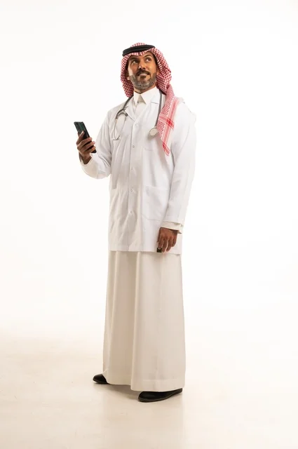Using a mobile phone for daily tasks, taking care of appearance, providing medical examinations and treatment services, a close-up portrait of a smiling Arab Gulf Saudi doctor wearing a doctor's coat and stethoscope, the concept of healthcare, white background.