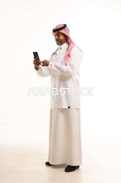 استخدام جهاز الهاتف المحمول في المهام اليومية، الاهتمام بالمظهر الخارجي، تقديم الفحص الطبي والخدمات العلاجية، بورتريه مقرب لطبيب عربي خليجي سعودي مبتسم يرتدي معطف الطبيب ويضع السماعات الطبية، مفهوم الرعاية الصحية، خلفية بيضاء