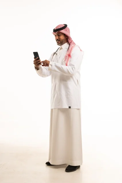 استخدام جهاز الهاتف المحمول في المهام اليومية، الاهتمام بالمظهر الخارجي، تقديم الفحص الطبي والخدمات العلاجية، بورتريه مقرب لطبيب عربي خليجي سعودي مبتسم يرتدي معطف الطبيب ويضع السماعات الطبية، مفهوم الرعاية الصحية، خلفية بيضاء
