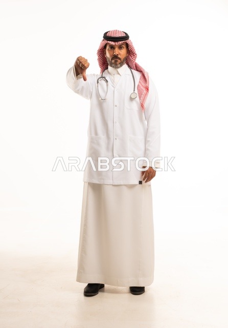 أشارة أصبع الأبهام للأسفل النظر إلى الكاميرا بايماءات الثقة بالنفس، الاهتمام بالمظهر الخارجي، تقديم الفحص الطبي والخدمات العلاجية، بورتريه مقرب لطبيب عربي خليجي سعودي مبتسم يرتدي معطف الطبيب ويضع السماعات الطبية، مفهوم الرعاية الصحية، خلفية بيضاء