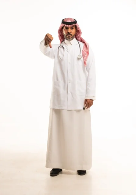 أشارة أصبع الأبهام للأسفل النظر إلى الكاميرا بايماءات الثقة بالنفس، الاهتمام بالمظهر الخارجي، تقديم الفحص الطبي والخدمات العلاجية، بورتريه مقرب لطبيب عربي خليجي سعودي مبتسم يرتدي معطف الطبيب ويضع السماعات الطبية، مفهوم الرعاية الصحية، خلفية بيضاء