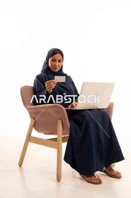 إنجاز مهام العمل عن بعد، التسوق الالكتروني وطلبات الاونلاين ، دردشة ومحادثات و تواصل اجتماعي ، اجراء اجتماع عمل عبر الانترنت، بورتريه لسيدة عربية سعودية خليجية ترتدي البالطو تجلس على كرسي مريح وتستخدم الحاسوب المحمول، خلفية بيضاء