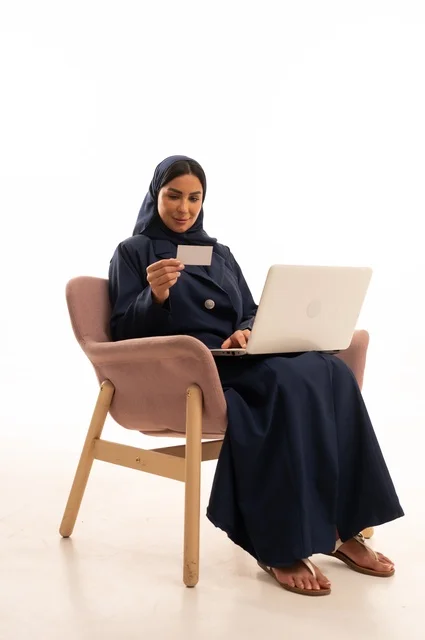 Completing remote work tasks, online shopping and orders, chatting and social communication, conducting a business meeting online, a portrait of a Saudi Arabian Gulf woman wearing a coat sitting on a comfortable chair and using a laptop, white background.