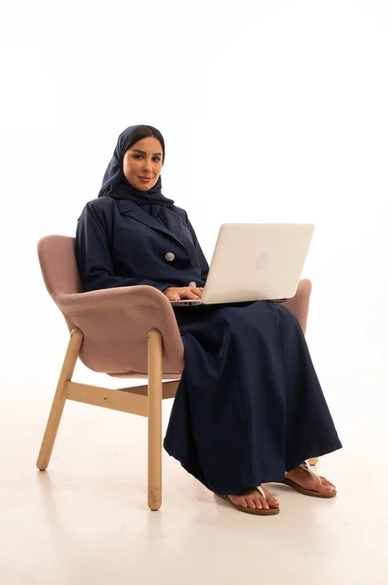 إنجاز مهام العمل عن بعد، التسوق الالكتروني وطلبات الاونلاين ، دردشة ومحادثات و تواصل اجتماعي ، اجراء اجتماع عمل عبر الانترنت، بورتريه لسيدة عربية سعودية خليجية ترتدي البالطو تجلس على كرسي مريح وتستخدم الحاسوب المحمول، خلفية بيضاء