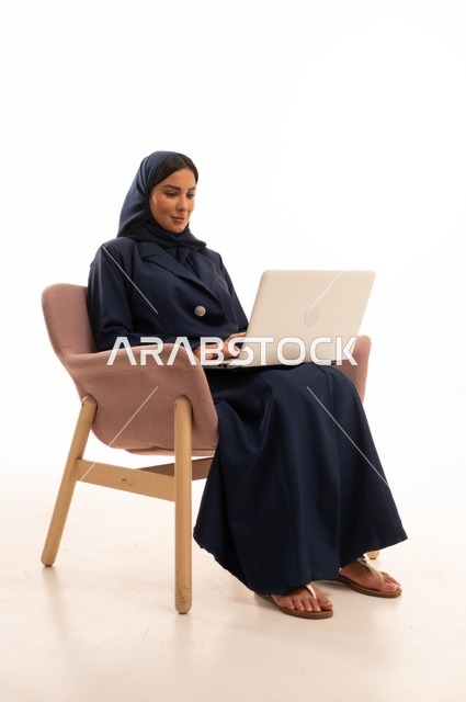 إنجاز مهام العمل عن بعد، التسوق الالكتروني وطلبات الاونلاين ، دردشة ومحادثات و تواصل اجتماعي ، اجراء اجتماع عمل عبر الانترنت، بورتريه لسيدة عربية سعودية خليجية ترتدي البالطو تجلس على كرسي مريح وتستخدم الحاسوب المحمول، خلفية بيضاء