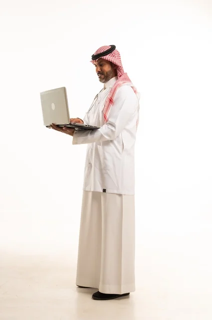 دمج التكنولوجيا مع الحياة العملية، الاهتمام بالمظهر الخارجي،  تقديم الفحص الطبي والخدمات العلاجية، بورتريه مقرب لطبيب عربي خليجي سعودي مبتسم يرتدي معطف الطبيب ويضع السماعات الطبية يحمل بيده حاب محمول يستخدمه لمراجعة حالات المرضى، الرعاية الصحية، خلفية بيضاء