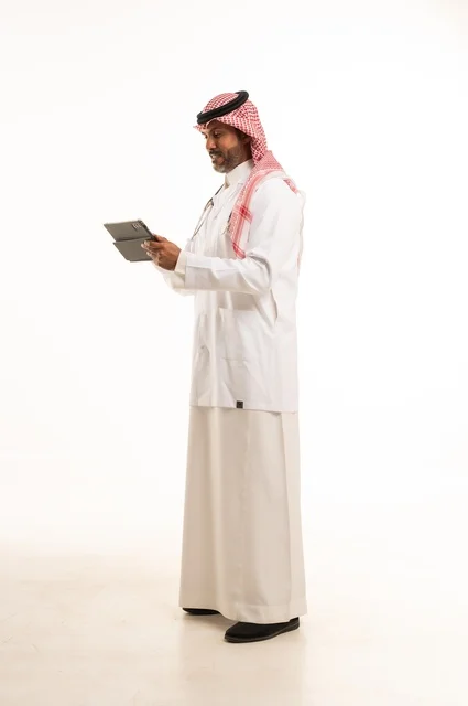 دمج التطور التقني في الحياة العملية، الاهتمام بالمظهر الخارجي، تقديم الفحص الطبي والخدمات العلاجية، بورتريه مقرب لطبيب عربي خليجي سعودي مبتسم يرتدي معطف الطبيب ويضع السماعات الطبية يحمل بيده جهاز لوحي يستخدمه لمراجعة حالات المرضى، الرعاية الصحية، خلفية بيضاء