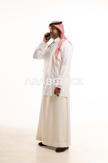 اجراء مكالمة هاتفية على الجهاز المحمول، الاهتمام بالمظهر الخارجي، تقديم الفحص الطبي والخدمات العلاجية، بورتريه مقرب لطبيب عربي خليجي سعودي مبتسم يرتدي معطف الطبيب ويضع السماعات الطبية، مفهوم الرعاية الصحية، خلفية بيضاء