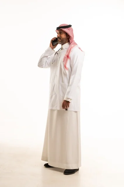 اجراء مكالمة هاتفية على الجهاز المحمول، الاهتمام بالمظهر الخارجي، تقديم الفحص الطبي والخدمات العلاجية، بورتريه مقرب لطبيب عربي خليجي سعودي مبتسم يرتدي معطف الطبيب ويضع السماعات الطبية، مفهوم الرعاية الصحية، خلفية بيضاء