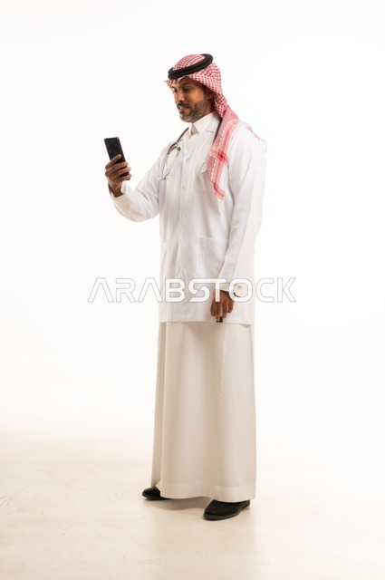 استخدام جهاز الهاتف المحمول في المهام اليومية، الاهتمام بالمظهر الخارجي، تقديم الفحص الطبي والخدمات العلاجية، بورتريه مقرب لطبيب عربي خليجي سعودي مبتسم يرتدي معطف الطبيب ويضع السماعات الطبية، مفهوم الرعاية الصحية، خلفية بيضاء