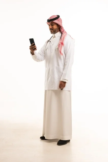 استخدام جهاز الهاتف المحمول في المهام اليومية، الاهتمام بالمظهر الخارجي، تقديم الفحص الطبي والخدمات العلاجية، بورتريه مقرب لطبيب عربي خليجي سعودي مبتسم يرتدي معطف الطبيب ويضع السماعات الطبية، مفهوم الرعاية الصحية، خلفية بيضاء